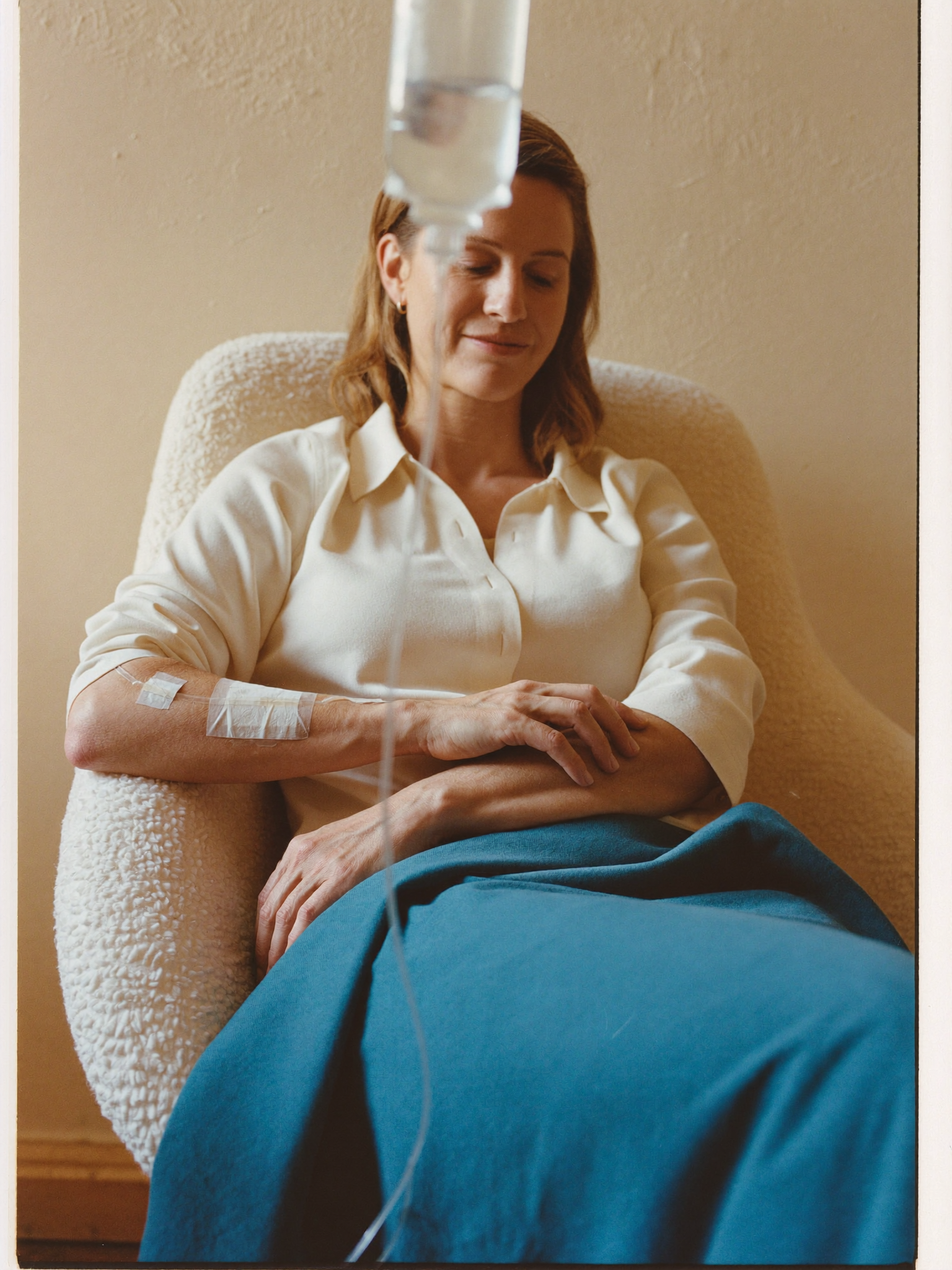 A woman relaxed in a cream chair with an IV line taped to her arm and a teal blanket over her lap